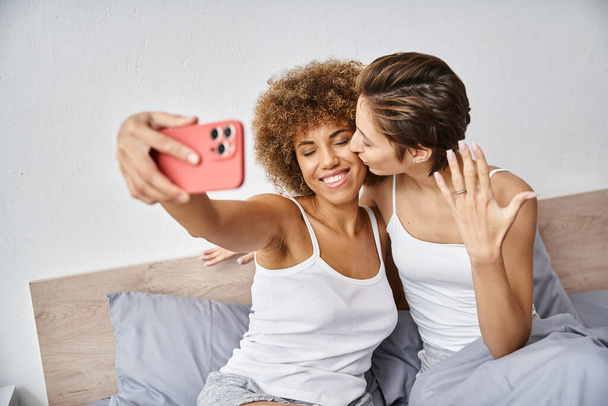 feliz y comprometido interracial lesbianas pareja tomando selfie en smartphone en casa, anillo de compromiso - Foto, Imagen