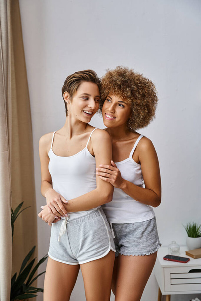 smiling african american woman with curly hair hugging girlfriend in pajamas and standing in bedroom - Photo, Image