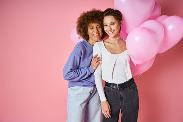 Valentines day concept, joyful multicultural lesbian couple smiling near heart shaped balloons - Photo, Image