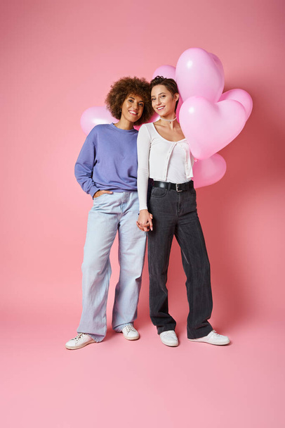 Valentines day concept, positive multicultural lesbian couple smiling near heart shaped balloons - 写真・画像