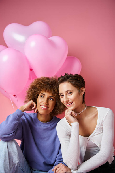 fröhliches multikulturelles lesbisches Paar, das neben rosafarbenen Herzballons sitzt, Valentinstag - Foto, Bild