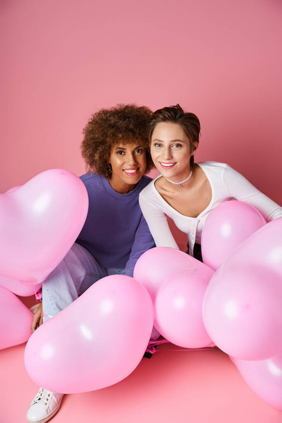 счастливая мультикультурная пара лесбиянок, сидящих вместе возле розовых шаров в форме сердца, День Святого Валентина - Фото, изображение