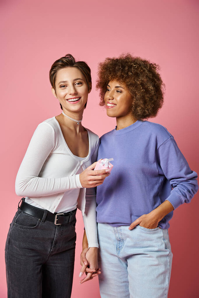 happy lesbian woman holding present and hand of african american girlfriend on Valentins day, pink - Фото, зображення