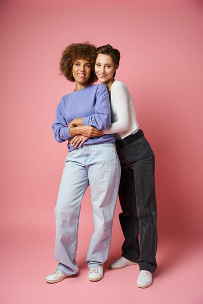 θετική λεσβία γυναίκα αγκαλιάζει αφροαμερικανή φίλη σε casual ρούχα σε ροζ φόντο - Φωτογραφία, εικόνα