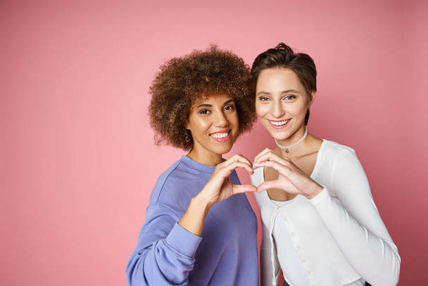 gioiosa multiculturale e lesbica coppia mostrando il segno del cuore con le mani su sfondo rosa, lgbtq amore - Foto, immagini