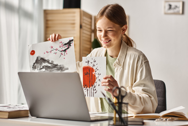 счастливая девочка-подросток, показывающая свои работы во время учебы и глядя на свой ноутбук, арт-класс в Интернете - Фото, изображение