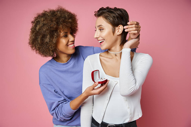 afro-amerikai nő holding box eljegyzési gyűrű közel meglepett barátnő rózsaszín háttér - Fotó, kép
