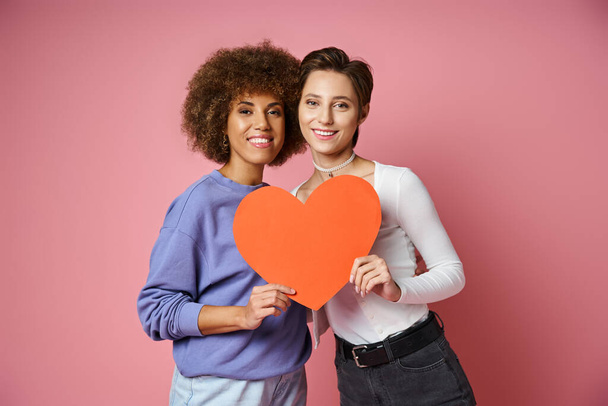 счастливая лесбийская и многонациональная пара, держащая красную коробку сердца, позируя на розовый, День Святого Валентина - Фото, изображение