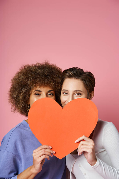 happy lesbian and multiethnic couple hiding behind red carton heart on pink, Valentines day - Photo, Image