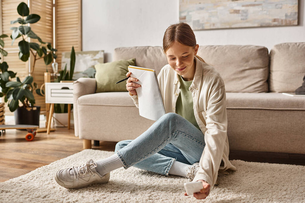 menina adolescente feliz segurando seu notebook e smartphone e sentado perto do sofá com fones de ouvido - Foto, Imagem