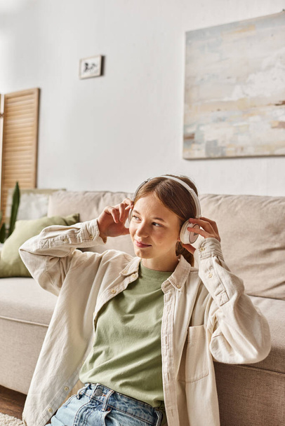 ανέμελη έφηβη κοπέλα χαλαρώνοντας με τα ασύρματα ακουστικά της και ακούγοντας μουσική κοντά στον καναπέ - Φωτογραφία, εικόνα