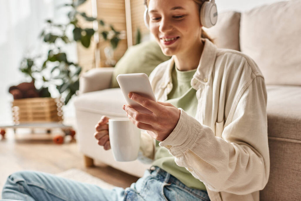 Kablosuz kulaklıklı gülümseyen genç kız evde çay içerken akıllı telefon kullanıyor. - Fotoğraf, Görsel