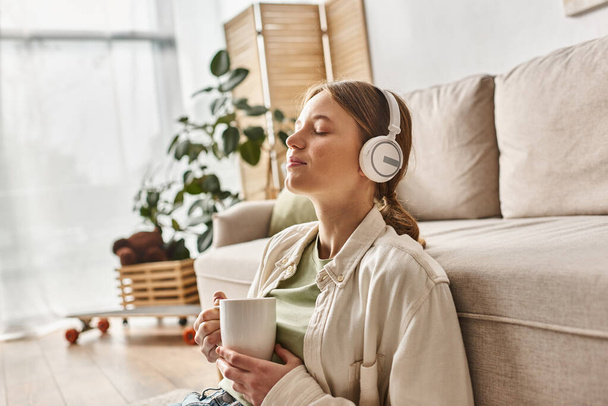 bezstarostný teenager dívka v bezdrátových sluchátkách poslech hudby a drží šálek čaje doma - Fotografie, Obrázek