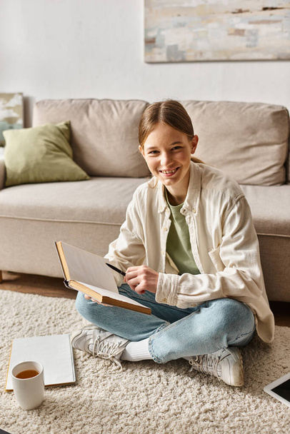 vrolijk tienermeisje lezen boek terwijl het doen van haar huiswerk en zitten op tapijt in de buurt van kopje thee - Foto, afbeelding