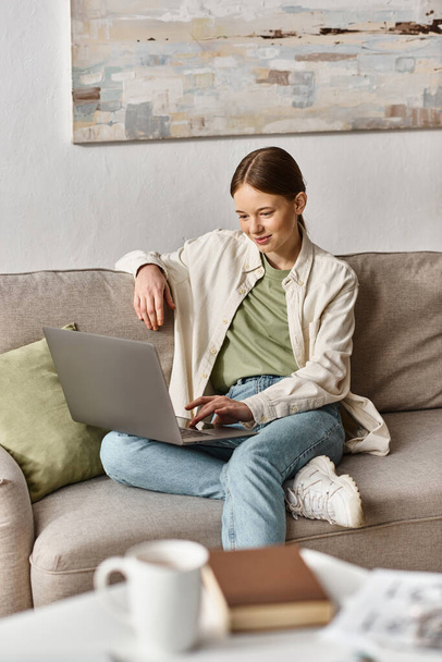 lächelndes Teenie-Mädchen, das sich auf Bildung konzentriert, indem es seinen Laptop benutzt und zu Hause auf einem bequemen Sofa sitzt - Foto, Bild