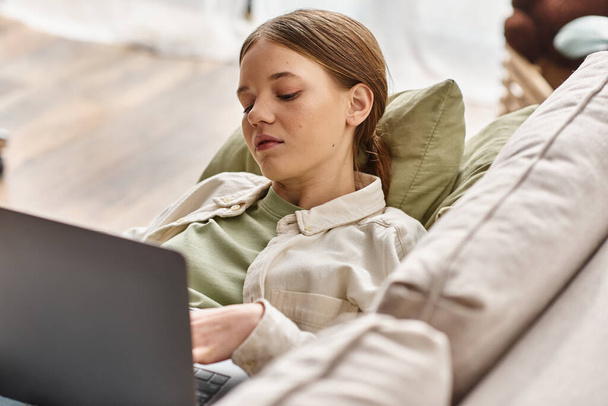 έφηβος κορίτσι επικεντρώθηκε στην e-learning χρησιμοποιώντας το laptop της και βρίσκεται σε ένα άνετο καναπέ στο σπίτι - Φωτογραφία, εικόνα