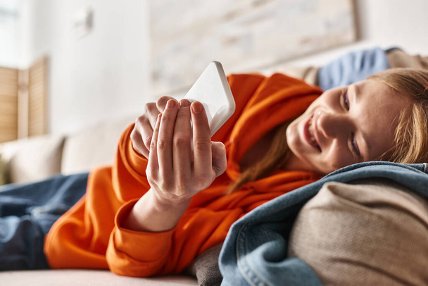 веселая девочка-подросток с помощью смартфона и сидя на диване в гостиной, пользователь социальных сетей - Фото, изображение