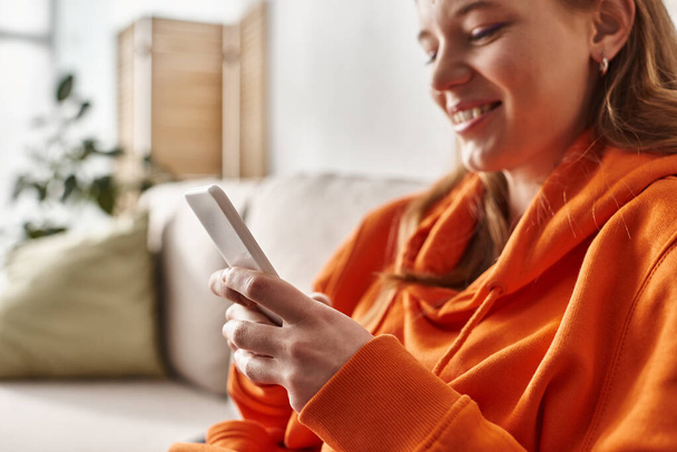 jolly teenage girl using her smartphone and sitting on sofa in living room, social media user - Zdjęcie, obraz