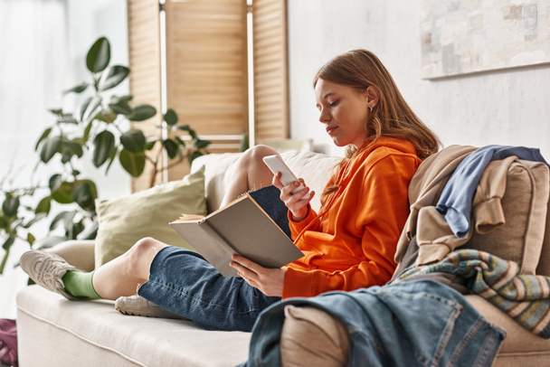 Teenager-Mädchen mit Buch und Smartphone auf Sofa neben chaotischem Kleiderstapel - Foto, Bild