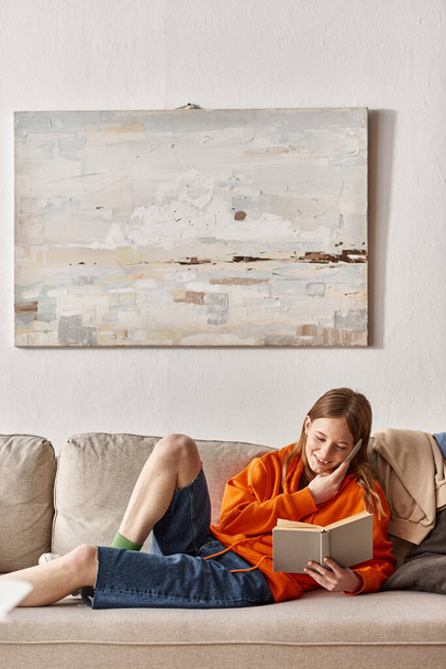 adolescente chica celebración de libro durante la llamada telefónica y sentado en el sofá al lado de un montón de ropa desordenado - Foto, imagen