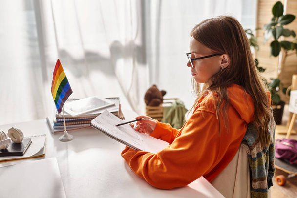стомлена дівчина-підліток в окулярах малює ескіз, занурена в творчий процес біля прапора гордості - Фото, зображення