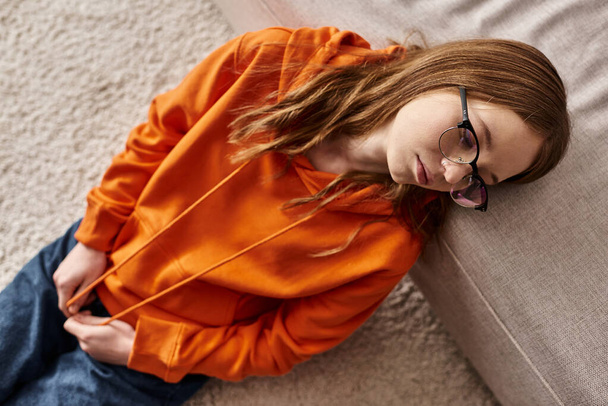 Draufsicht auf Mädchen in orangefarbenen Kapuzenpullovern in der Nähe des heimischen Sofas, Melancholie und Einsamkeit eines Teenagers - Foto, Bild