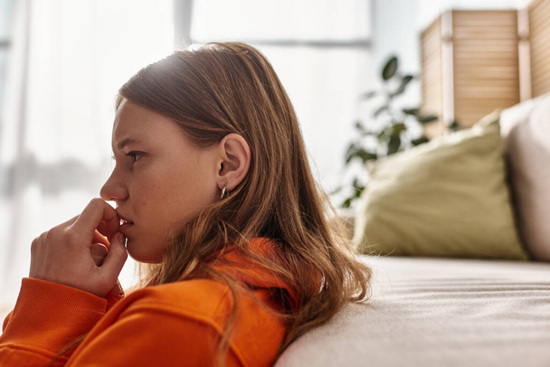 side view of teenage girl in distress looking away while sitting near couch in living room - Valokuva, kuva