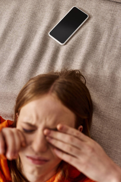 вид сверху на девочку-подростка в оранжевой толстовке, лежащую рядом со смартфоном на диване, стресс от социальных сетей - Фото, изображение