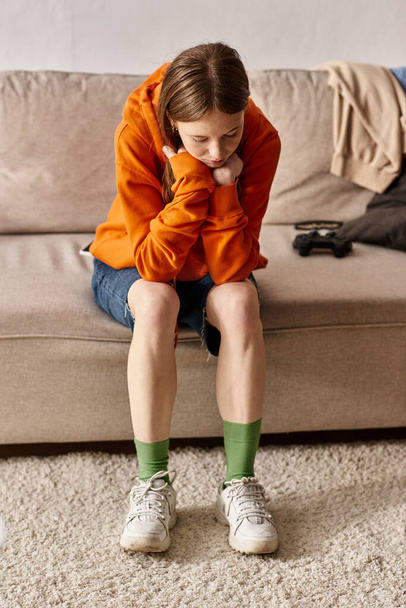upset teenager girl in orange hoodie sitting on couch near blurred joystick, solitude and sadness - Photo, Image