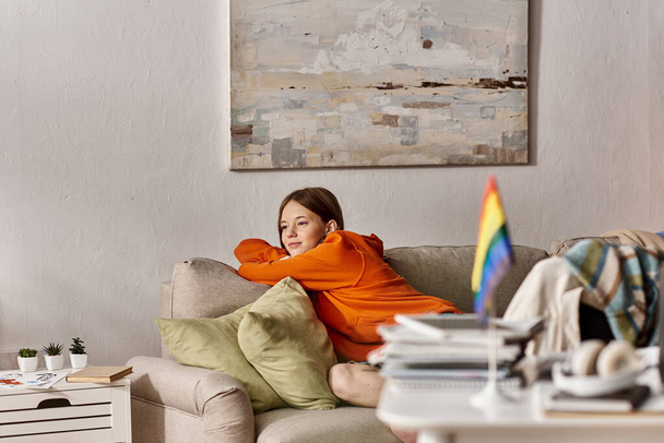 verträumtes Teenie-Mädchen im Kapuzenpulli sitzt auf Couch mit distanziertem Blick, verschwommene lgbtq-Flagge im Vordergrund - Foto, Bild