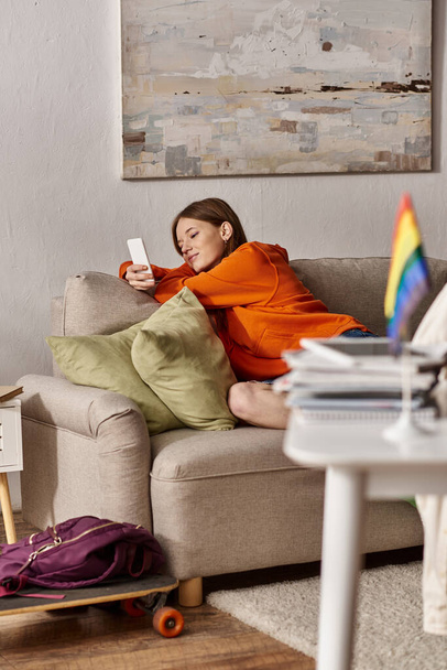 Alegre chica adolescente usando su teléfono inteligente cerca del sofá y borrosa bandera lgbt en primer plano - Foto, imagen