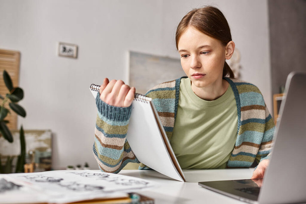 Skoncentrowany nastolatek dziewczyna patrząc na notatnik podczas e-learning w pobliżu laptopa i szkice na biurku - Zdjęcie, obraz