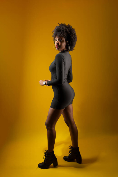 Sensual young woman standing, wearing black clothes and posing for a photo. Isolated on yellow background. - Fotoğraf, Görsel