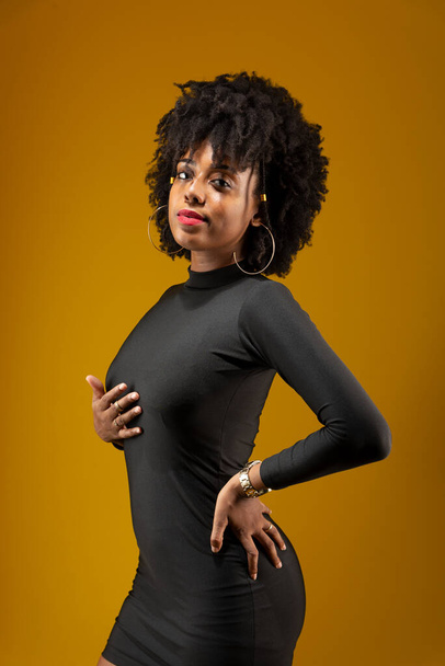 Mujer joven y sensual, de pelo negro, de pie, vestida de negro y posando para una foto. Aislado sobre fondo amarillo. - Foto, imagen