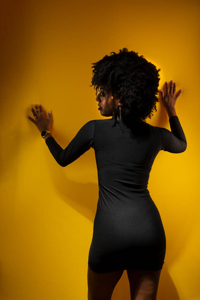 Young sensual woman with black power hair, wearing black clothes leaning against a yellow wall posing for a photo. - Foto, Imagen