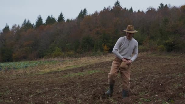 Vetés a vetőmag a betakarítás: Farmer a Country Attire Séták át szántott területen alatt őszi égbolt. Kiváló minőségű 4k felvétel. Vidéki ruhába és kalapba öltözött parasztként rusztikus útra indul. - Felvétel, videó