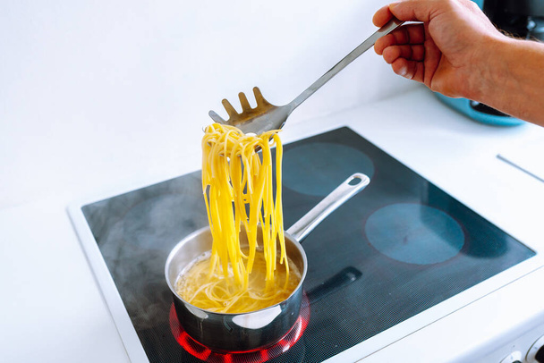 mężczyzna gotuje makaron spaghetti w domu w kuchni. mężczyzna gotuje w brązowym fartuchu, próbuje spaghetti gotowość, trzyma spaghetti na specjalnej łyżce nad wrzącą wodą. Koncepcja włoskiej kuchni domowej - Zdjęcie, obraz