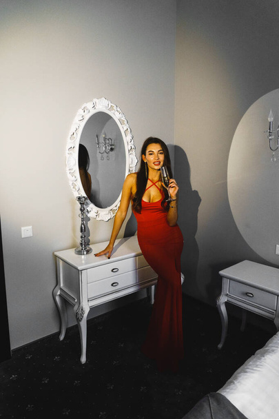 Femme sexy avec une coupe de champagne dans une robe rouge posant dans une chambre d'hôtel. Concept de vacances coûteux - Photo, image