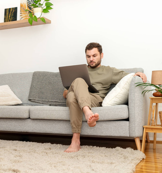 Mann sitzt zu Hause auf Sofa und benutzt Laptop. Arbeiten von zu Hause aus. Innenbehörde. - Foto, Bild