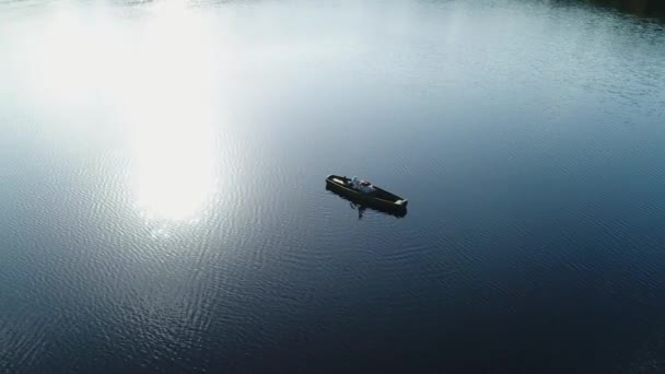 Mies soittaa kitaraa makatessaan puuveneessä keskellä villiä järveä veden pinnalla auringon säteiden heijastuessa aaltoihin. Antenninäköala. - Materiaali, video