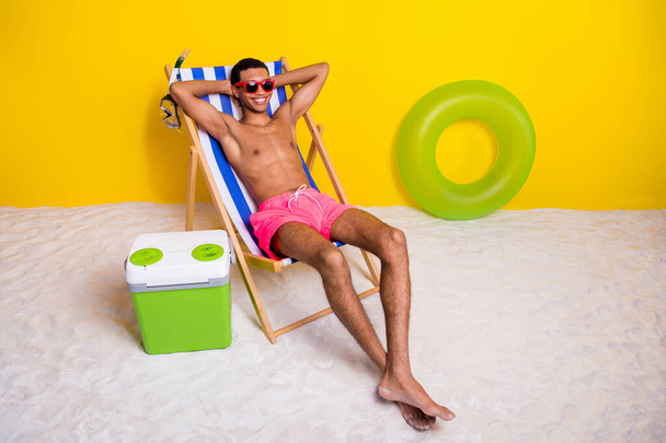 Full length photo of cheerful funky shirtless man relaxing lounge chair arms behind head isolated yellow color background. - 写真・画像
