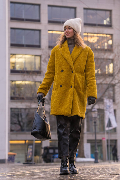 Κάτω όψη μιας γυναίκας που περπατάει στη μέση ενός άδειου δρόμου της πόλης. Η κυρία φοράει ένα κίτρινο παλτό. Κοιτάζει την πόλη και ενδιαφέροντα μέρη.. - Φωτογραφία, εικόνα