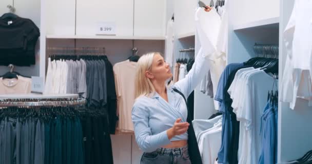 Woman shopping in clothing boutique. Smiling happy stylish trendy woman with great sense of style chooses seasonal trendy clothes, creates unique outfits. Woman evaluating material quality of t-shirt - Footage, Video