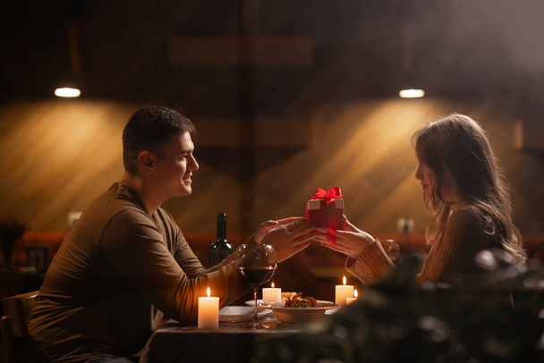 Feliz pareja joven celebrando el día de San Valentín teniendo una cena romántica en casa. Hombre cariñoso dando caja de regalo a su mujer. Copiar espacio - Foto, Imagen
