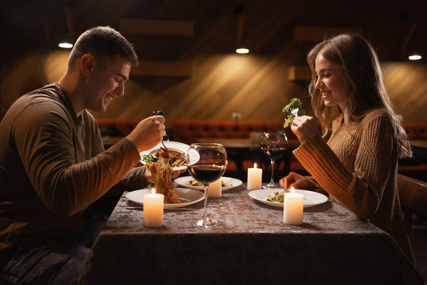 νεαρό ζευγάρι ερωτευμένο να δειπνεί σε ένα εστιατόριο γιορτάζοντας την ημέρα του Αγίου Βαλεντίνου, ο άντρας να βάζει ζυμαρικά σε ένα πιάτο και η γυναίκα να χαμογελάει. Ρομαντικό δείπνο έννοια - Φωτογραφία, εικόνα