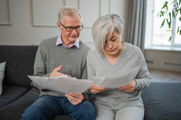Sad tired disappointed middle aged senior couple sit with paper document. Unhappy older mature man woman reading paper bill managing bank finances calculating taxes planning loan debt pension payment - Photo, Image