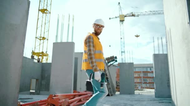 Gelukkige bouwvakker op bouwplaats dansend met boxtool en schroevendraaier. Geniet van het moment dat hij uniform en veiligheidshelm draagt. Kijkend naar camera - Video