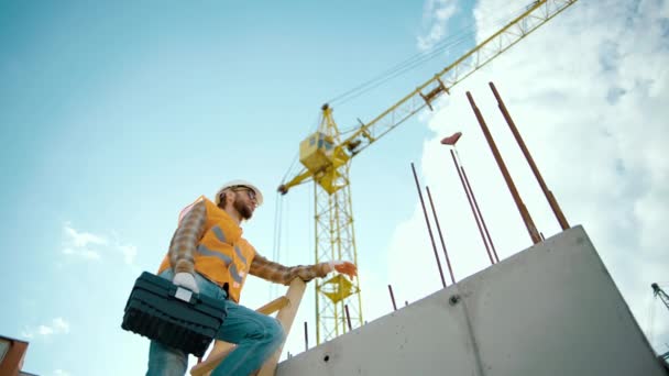 Professionelle Bauarbeiter erklimmen hohe Holzleiter mit Werkzeugkiste in der Hand. Bau und Kran im Hintergrund - Filmmaterial, Video