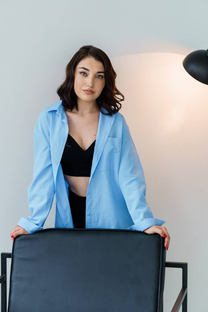 Beautiful girl in black trousers and a blue jacket near a chair on a white background. Portrait of a beautiful young woman - Φωτογραφία, εικόνα