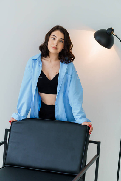 Siyah pantolonlu güzel bir kız ve beyaz arka planda bir sandalyenin yanında mavi bir ceket. Güzel bir genç kadının portresi. - Fotoğraf, Görsel